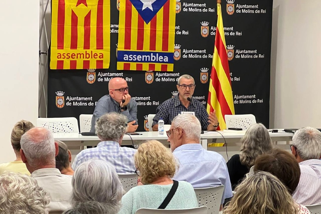 A la imatge es veu una taula on parlen dos dels membres de l'ACN a través de uns micros, davant una nombrosa audiència. La part del darrere té unes banderes penjades amb la senyera catalana i una banda on posa "assemblea".